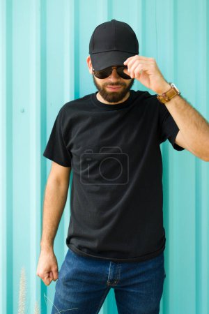 Téléchargez les photos : Homme hispanique à la mode dans un t-shirt et une casquette noirs tient sa casquette avec désinvolture, dans un contexte turquoise, parfait pour une maquette - en image libre de droit