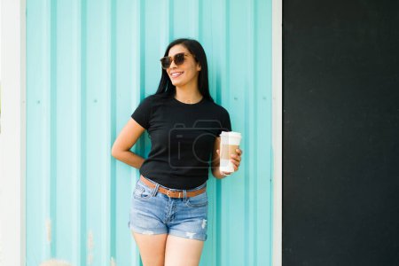 Téléchargez les photos : Femme hispanique souriante en t-shirt noir tenant une tasse de café se tient avec confiance sur un fond sarcelle avec des lunettes de soleil par une journée ensoleillée - en image libre de droit