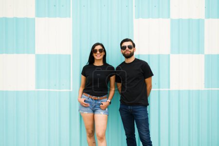 Foto de Pareja hispana segura con gafas de sol posando sobre un fondo de rayas turquesas y blancas, con espacio para el logo en sus camisetas negras - Imagen libre de derechos