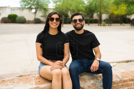 Foto de Sonriente pareja hispana joven con camisetas negras en blanco y gafas de sol posando para una maqueta en un entorno exterior brillante - Imagen libre de derechos
