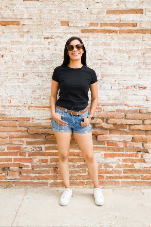 Téléchargez les photos : Femme latina auto-assurée posant devant un mur de briques, présentant un simple t-shirt noir idéal pour les maquettes - en image libre de droit