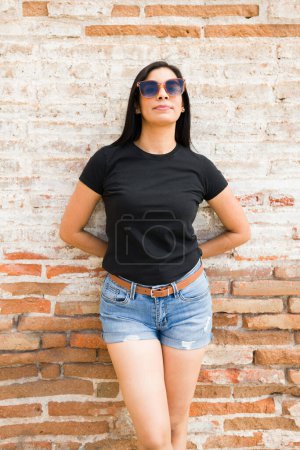 Téléchargez les photos : Femme hispanique élégante portant un t-shirt noir uni et un short en denim pose en toute confiance sur un fond mural en brique rustique - en image libre de droit