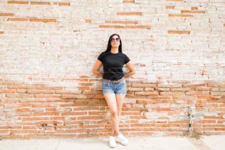 Téléchargez les photos : Jolie femme latine en t-shirt noir et short en denim, pose pleine longueur avec des lunettes de soleil devant un mur de briques texturées, idéale pour les maquettes - en image libre de droit