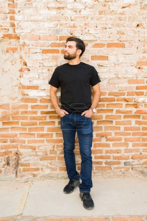 Foto de Retrato completo del hombre hispano que exuda confianza en una camiseta negra en blanco para una presentación de maqueta, posando frente a un fondo rústico de pared de ladrillo - Imagen libre de derechos