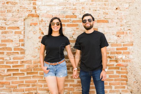 Foto de Alegre pareja hispana cogida de la mano, con camisetas negras en blanco adecuadas para maquetas de marca, de pie ante una vieja pared de ladrillo - Imagen libre de derechos