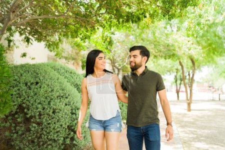 Téléchargez les photos : Jeune couple latin souriant et marchant ensemble à l'extérieur, profitant d'une journée ensoleillée lors d'un rendez-vous romantique décontracté dans le parc - en image libre de droit