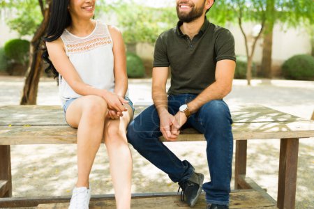 Photo for Cheerful young couple enjoying a sunny day on a bench outdoors, sharing smiles and conversation, giving off a relaxed latin romance atmosphere - Royalty Free Image