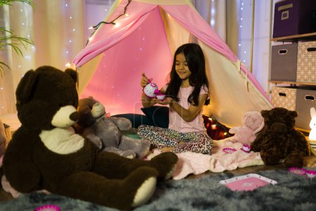 Téléchargez les photos : Petite fille hispanique dégustant un thé entouré d'animaux en peluche dans une tente de chambre confortable avec des veilleuses magiques - en image libre de droit