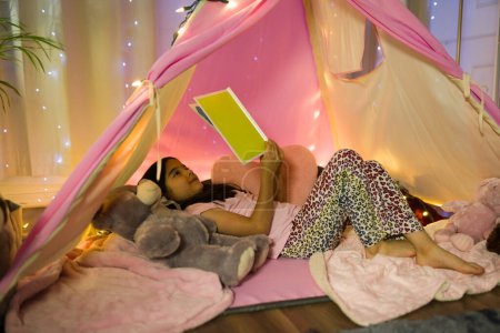 Foto de Joven chica latina disfruta leyendo un libro en una tienda casera en su dormitorio con luces de hadas por la noche, sintiéndose cómoda y segura - Imagen libre de derechos