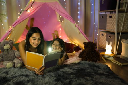 A heartwarming scene of a hispanic mother reading a bedtime story to her daughter in a cozy home bedroom teepee at night