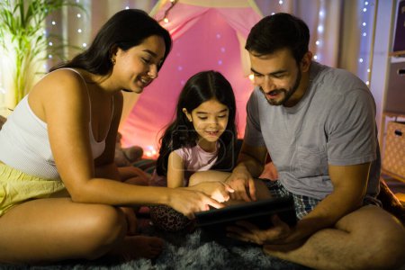 Foto de La familia disfruta de un momento cálido e íntimo juntos por la noche dentro de una tienda de campaña con luces de hadas, jugando con una tableta - Imagen libre de derechos