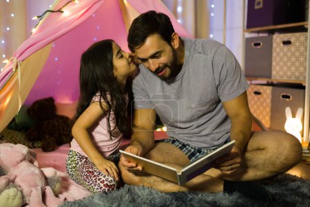 Vater liest seiner Tochter eine Gute-Nacht-Geschichte vor und hört einem Geheimnis zu, während sie ihm ins Ohr flüstert