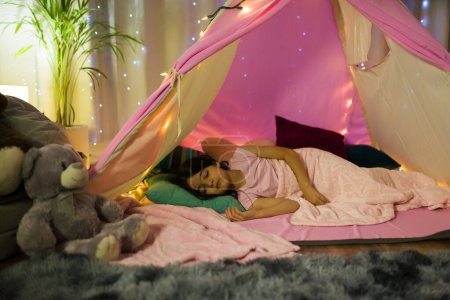 Foto de Pequeña chica latina que duerme tranquilamente en casa en un acogedor dormitorio con una caprichosa tienda de campaña y luces de estrellas brillantes por la noche - Imagen libre de derechos