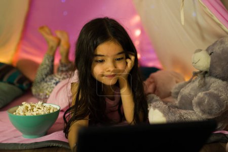 Téléchargez les photos : Petite fille couchée dans une tente, regardant un film sur sa tablette, avec du pop-corn et des jouets en peluche, dans une chambre à coucher - en image libre de droit