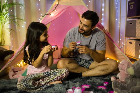 Foto de Padre e hija disfrutan de una fiesta de té juguetona juntos en una tienda casera, rodeados de luces de hadas, por la noche - Imagen libre de derechos