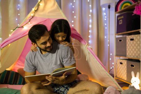 Photo for Father and his little girl share a bedtime story in a cozy homemade tent decorated with fairy lights, establishing a heartwarming nightly tradition - Royalty Free Image