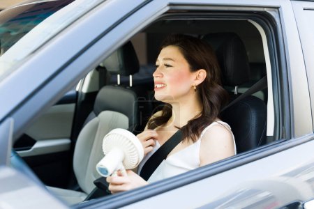 Photo for Stressed woman driving a car in the summer and suffering from the heat while holding a portable fan - Royalty Free Image