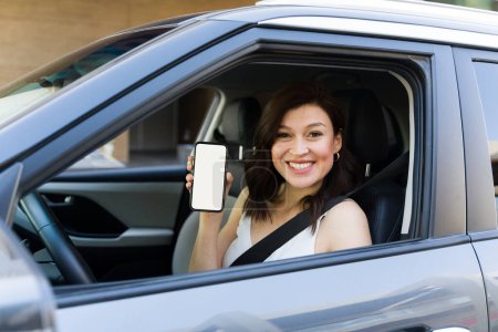 Schöne und fröhliche Fahrerin zeigt leeren Smartphone-Bildschirm, während sie in ihrem Auto sitzt, bereit für eine Fahrt
