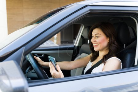 Foto de Mujer feliz en sus 30 años multitarea mediante el uso de un teléfono inteligente mientras conduce su coche, destacando los riesgos de conducción distraída - Imagen libre de derechos