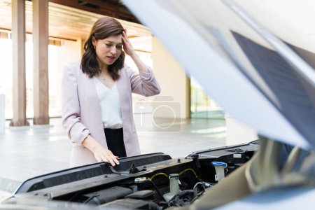 Téléchargez les photos : Inquiète jeune femme en tenue d'affaires examinant son moteur de voiture, se sentant frustré par la panne soudaine du véhicule dans le stationnement - en image libre de droit