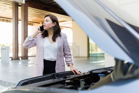 Concerned businesswoman on a call for assistance with an open car hood in a parking structure, facing potential vehicle issues