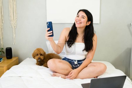 Photo for Happy young woman using smartphone to capture a selfie moment while sitting with her cute dog on a bed in a cozy bedroom - Royalty Free Image