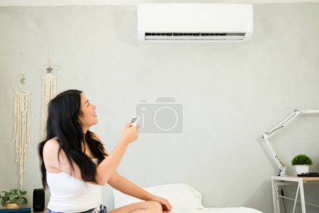 Téléchargez les photos : Caucasian woman adjusting a mini split air conditioning unit with a remote control while sitting comfortably on her bed on a hot summer day - en image libre de droit