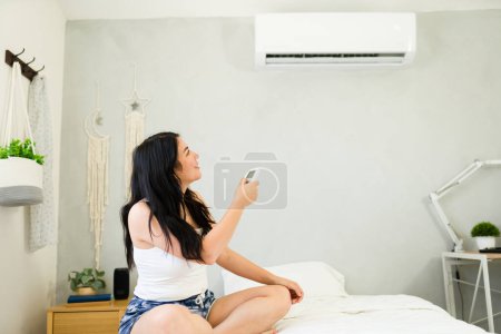 Téléchargez les photos : Young woman relaxes on her bed with a remote in hand, enjoying the cool summer atmosphere provided by the mini split air conditioning unit - en image libre de droit