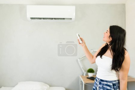 Téléchargez les photos : Cheerful woman stands by the bed, adjusting a mini split air conditioning unit with a remote control during a hot summer day - en image libre de droit