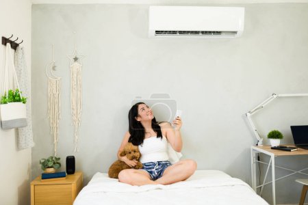 Téléchargez les photos : Smiling woman with a remote control enjoys the cooling breeze from a mini split air conditioning unit on a hot summer day - en image libre de droit
