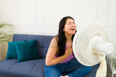 Caucasian woman seeking relief from the summer heat by sitting in front of a fan, visibly perspiring and trying to cool down at home