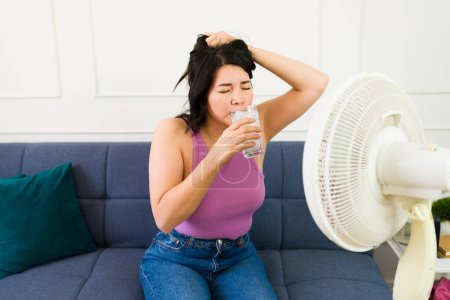 Caucasian woman finding relief from the summer heat at home with a fan, ice water, and relaxed indoor ambiance