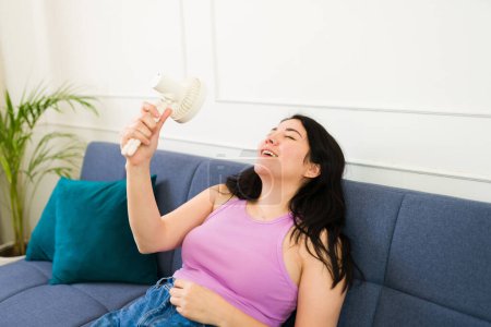 Beautiful woman sitting on her couch at home uses a portable fan to beat the sweltering summer heat 