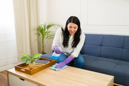 Photo for Caucasian woman in rubber gloves cleaning a wooden coffee table in a beautifully decorated and well-lit living room at home - Royalty Free Image