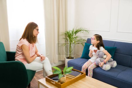 Photo for Little girl with a teddy bear talking to a female therapist during therapy session in a comfortable office, promoting mental health and support - Royalty Free Image