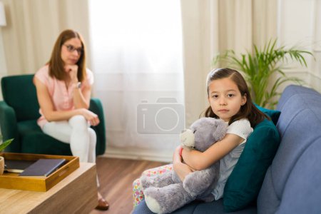 Photo for Cute little girl making eye contact while hugging a teddy bear during a therapy session with a child psychologist - Royalty Free Image