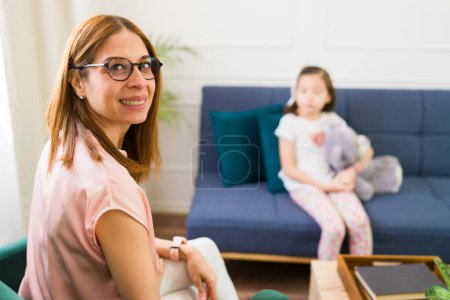Photo for Professional child psychologist smiling at the camera during a therapy session with a focused little girl sitting with a stuffed toy - Royalty Free Image