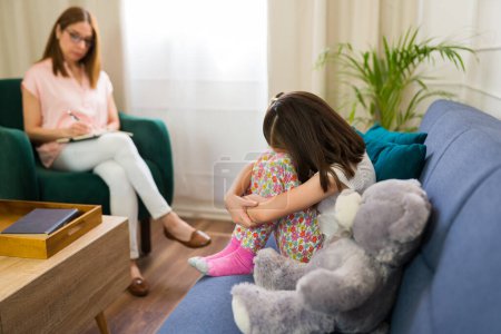 Photo for Little girl sits embracing her knees during a therapy session, with a child psychologist attentively observing in the background - Royalty Free Image