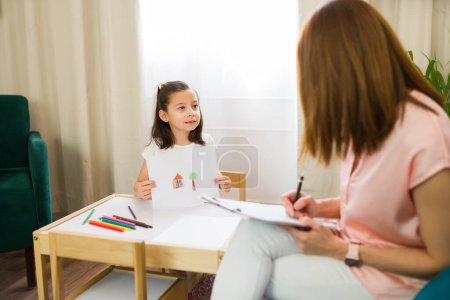 Concentrated child psychologist engages with a young girl as she shares her artwork during a therapeutic counseling session in a comfortable office setting