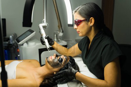 Woman receiving a YAG laser carbon mask facial treatment from an experienced beautician in a contemporary medical beauty clinic environment