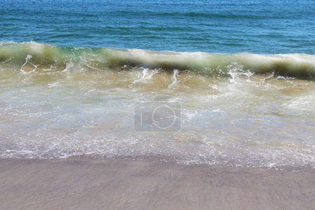 A wave is seen rolling into the shore of a beach, creating a dynamic and powerful scene. The water is foamy as it crashes onto the sandy shore, leaving behind a trail of white bubbles.
