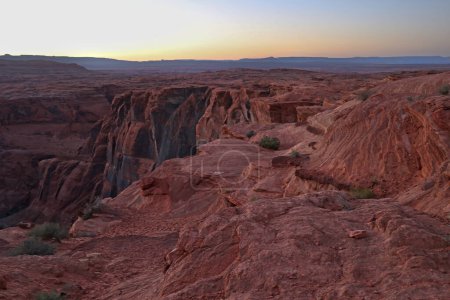 The sun slowly lowers on the horizon, casting a warm glow over the rugged desert canyon. The orange and red hues of the sky contrast with the dark shadows of the canyon walls.
