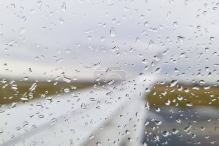 Rain drops have accumulated on the windshield of a stationary car, distorting the view outside. The water droplets create a pattern on the glass as they slide down, while the wipers remain motionless.
