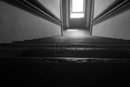 Schwarz-Weiß-Foto mit dunkler Treppe, die zu einem hell erleuchteten Fenster führt. die Perspektive von unten betont den dramatischen Kontrast und hebt die architektonischen Elemente und Texturen des Raumes hervor.