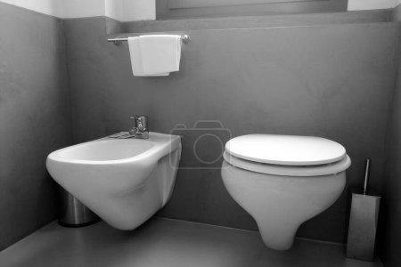 Monochrome Fotografie, die ein modernes minimalistisches Badezimmerdesign mit einer wandmontierten Toilette und einem Bidet nebeneinander zeigt. das elegante Design verfügt über eine saubere, neutrale Farbpalette, minimalistische Armaturen und ein Handtuch, das fein säuberlich auf einem Handtuchhalter gefaltet ist.