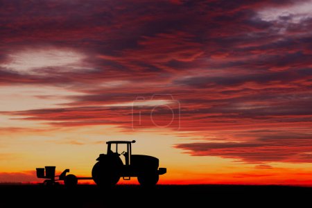 Silhouette of a tractor with a seeder against a bright sunset sky with dramatic clouds, depicting rural life and farming.
