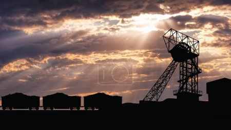 Silhouette of coal mine buildings and loaded freight train cars against a dramatic sunset sky with clouds and sun rays.