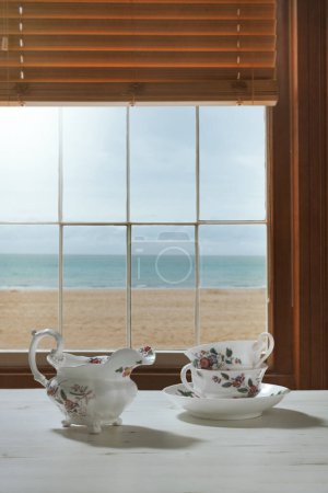 Foto de Tazas de té vintage y jarra de leche en la ventana con vistas al océano - Imagen libre de derechos