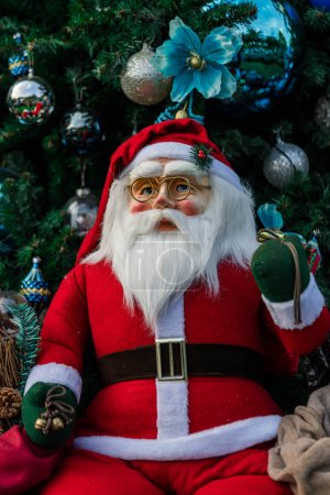 Téléchargez les photos : Poupée Santa Clause décorée devant le sapin de Noël avec des jouets ornementaux colorés lors du festival de Noël dans le monde entier - en image libre de droit