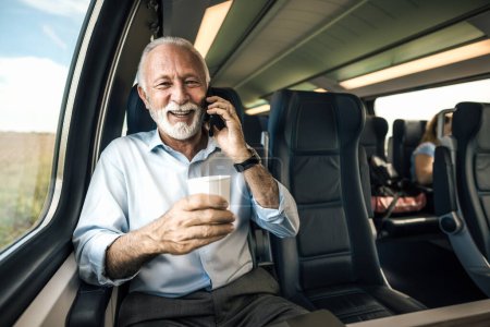 Photo for Smiling senior businessman talking on smart phone. Male entrepreneur is having coffee while sitting on subway train. He is wearing formals during business travel. - Royalty Free Image